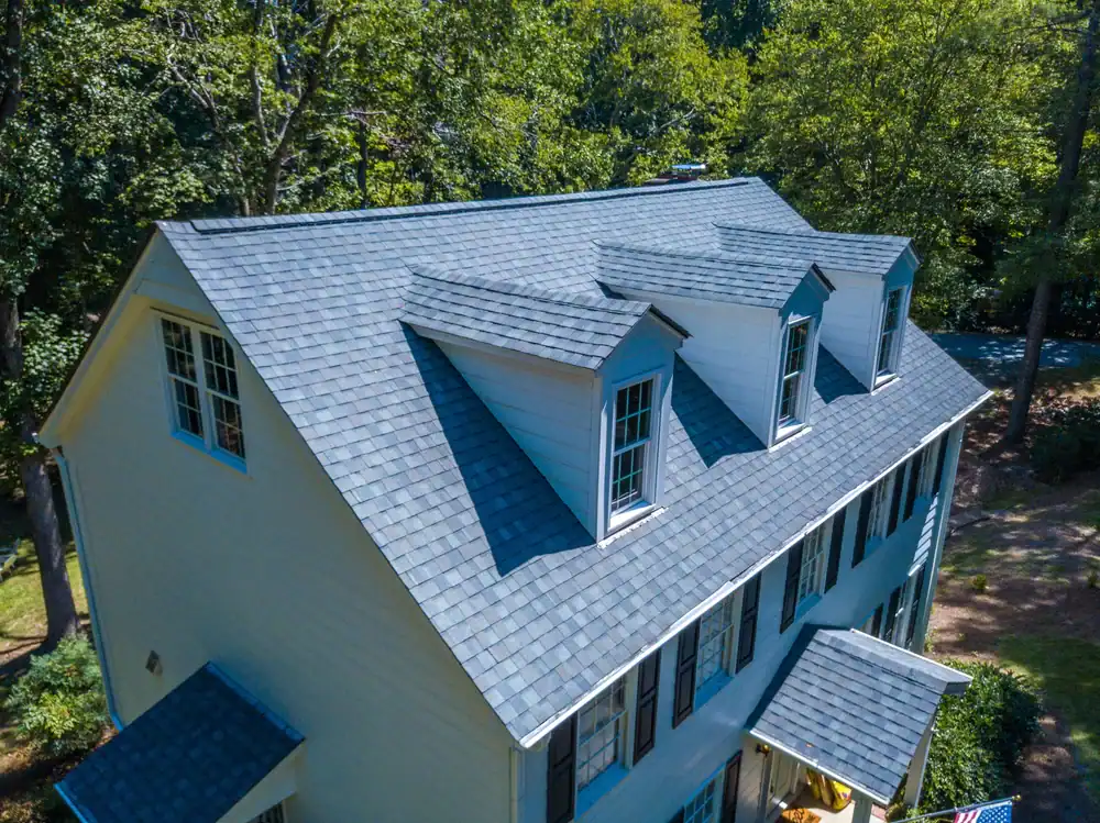 Aerial view of a house with a gray shingle roof featuring three dormer windows; the house, surrounded by trees and boasting white exterior walls with dark shutters, showcases work by a skilled roofing contractor in Southwest Louisiana, TX.