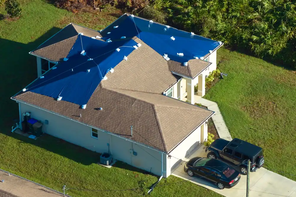 A house with tan-colored roofing is partially covered by a blue tarp, likely for storm protection. Two cars are parked in the driveway, and green grass and trees surround the home&mdash;a roofer Jasper County, TX may be needed for repairs.