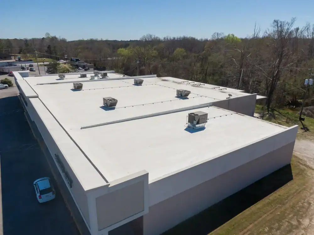 Aerial view of a large commercial building with a flat white roof, multiple HVAC units on top, and a single parked car by the side—ideal for a roofing contractor Southwest Louisiana project, surrounded by trees and clear skies.