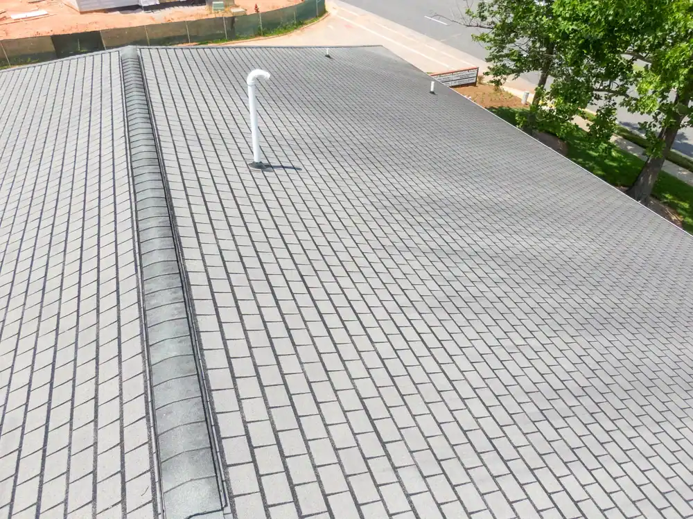 Aerial view of a clean, gray shingle roof with a white rooftop vent, expertly installed by a roofing contractor Southwest Louisiana. Trees, a street, and a construction site with dirt and fencing are visible in the background.