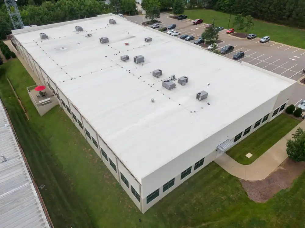 Aerial view of a large, white, flat-roofed commercial building in TX with multiple rooftop HVAC units, surrounded by grass, trees, and a parking lot with parked cars&mdash;ideal for a roofing contractor Southwest Louisiana project.