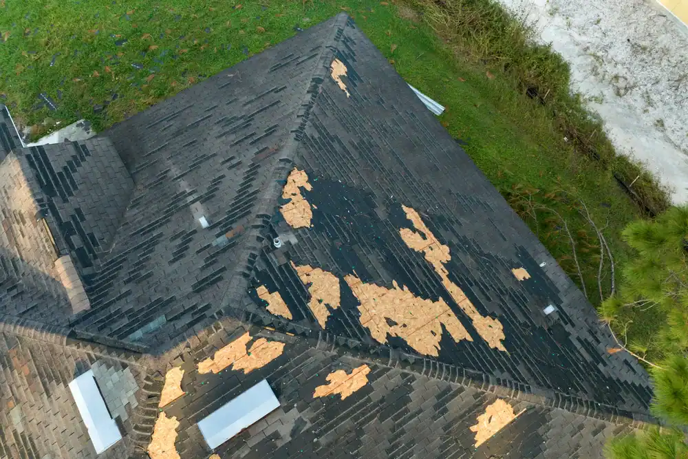 Aerial view of a house roof with significant shingle damage; many shingles are missing, exposing the underlying wood. Surrounding area includes green grass and some bushes&mdash;ideal for a roofing contractor Southwest Louisiana to assess and repair.