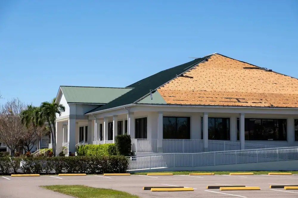 A building with a partially stripped roof, exposing wooden panels, stands under a clear blue sky. Nearby palm trees and bushes frame an empty parking lot&mdash;a typical scene for a roofing contractor Southwest Louisiana,TX project.
