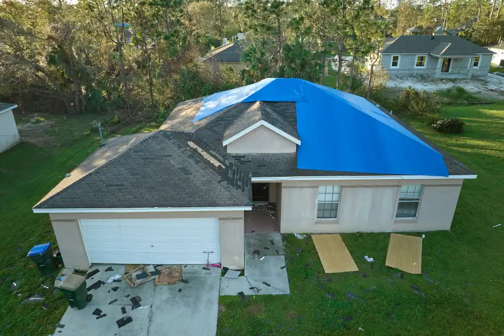A single-story house with a blue tarp covering part of its roof suggests repairs in progress by a roofing contractor Southwest Louisiana. Debris and wooden boards scatter the driveway and grass, surrounded by trees and neighboring homes.