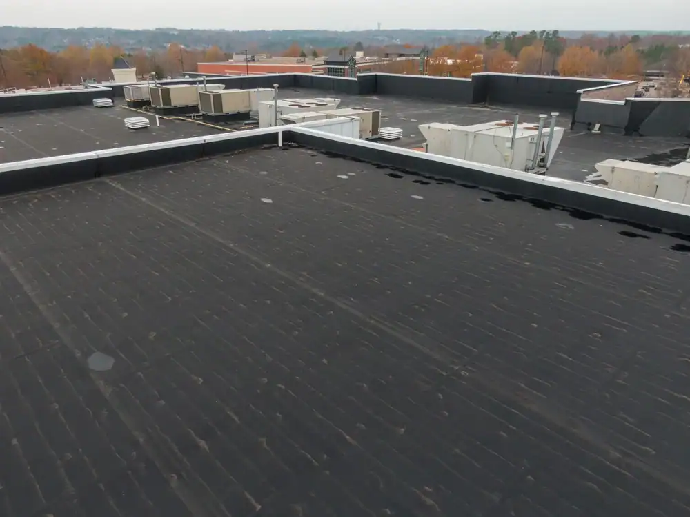 A flat commercial building roof with a black membrane surface, HVAC units, and other equipment, surrounded by a low parapet wall. A roofer Jasper County or roofing contractor Southwest Louisiana would recognize the expansive view of trees and nearby buildings.