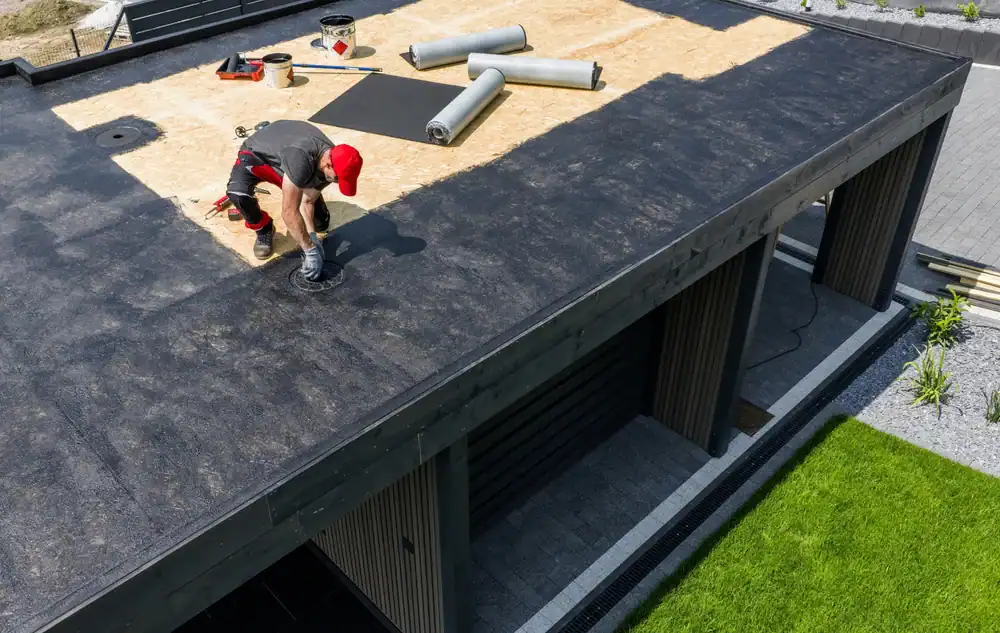 A roofer Jasper County, TX, wearing a red cap and gloves installs roofing material on a flat-roofed building, with rolls of roofing felt and tools scattered around. The building overlooks a green lawn and paved pathway.