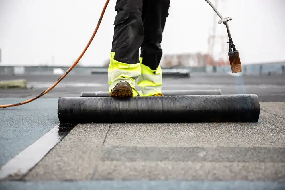 A roofer Jasper County in yellow safety pants unrolls roofing material on a flat roof, using a torch to seal it during roof construction or repair.