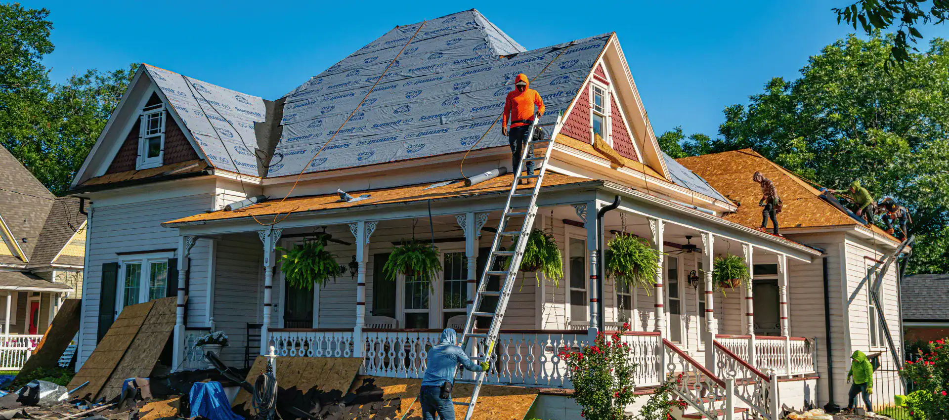 Workers replace the roof of a large house, with some standing on ladders and others on the roof. This TX home features white siding, a porch with plants, and roofing materials scattered around—skilled roofers at work in Jasper County.