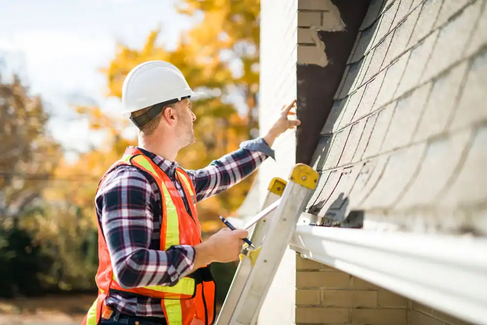 A roofing contractor Southwest Louisiana in a hard hat and safety vest stands on a ladder inspecting the exterior wall and roof shingles of a house on a sunny day, holding a clipboard and pen.