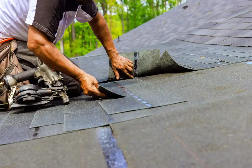 A roofer in Jasper County installs dark asphalt shingles on a sloped roof, using a tool to secure them. A nail gun rests nearby, while green trees frame the background.