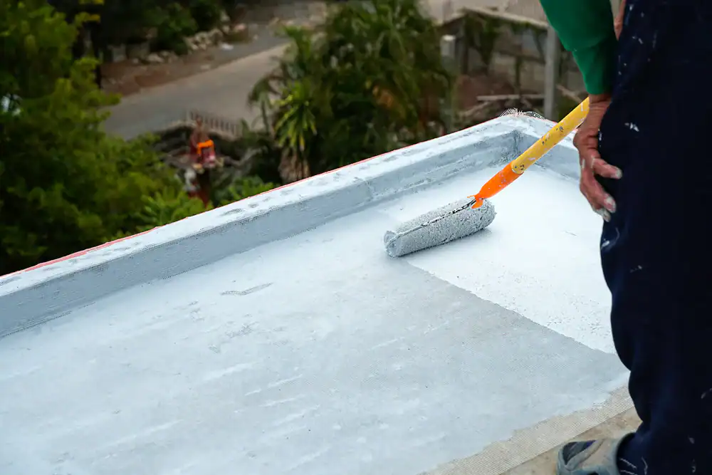 A roofer from Jasper County uses a paint roller to apply a white waterproofing coating to a flat concrete rooftop, with greenery and a street visible in the background.