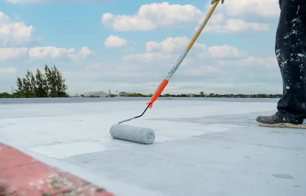 A person uses a paint roller with a long handle to apply white paint or coating to a flat rooftop under a partly cloudy sky. Only part of the person's legs and shoes are visible.