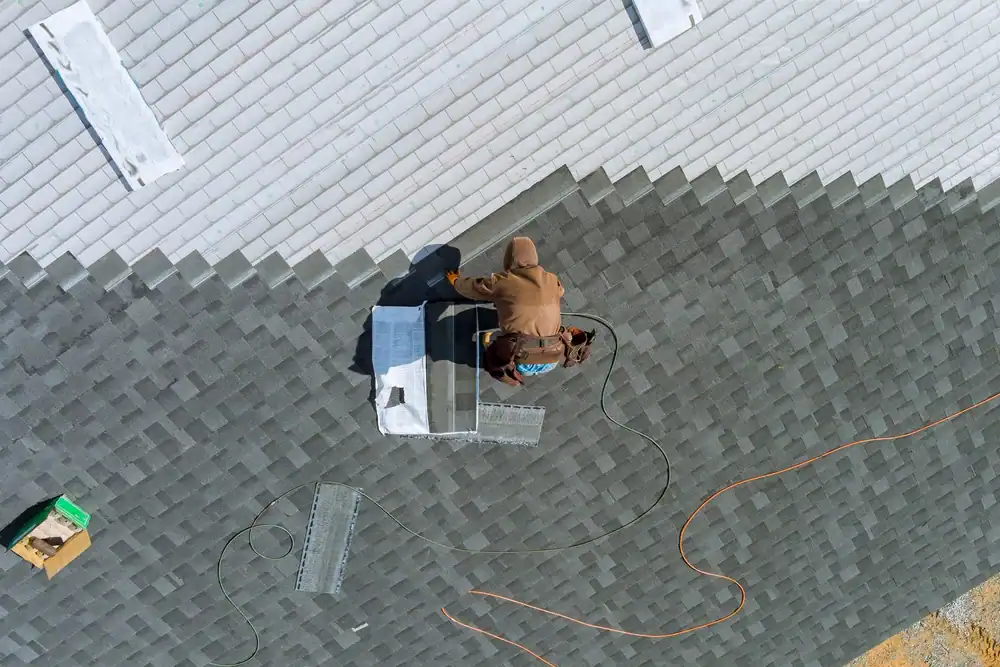 A roofing contractor Southwest Louisiana works on a shingled roof, using tools and materials spread around them. An orange extension cord and metal vents are visible near the worker on the rooftop.
