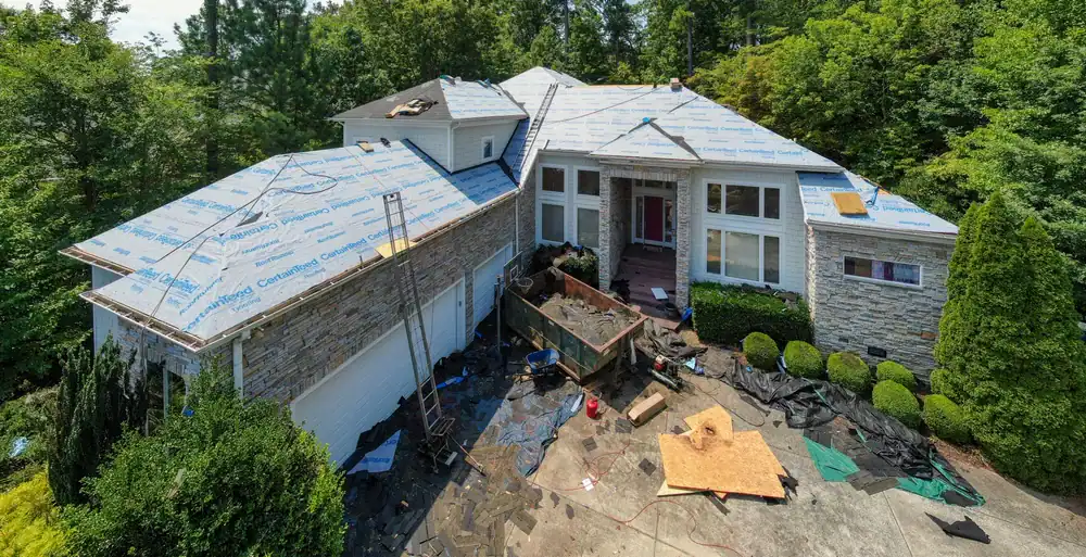 A large house is having its roof replaced by a roofing contractor Southwest Louisiana,TX; workers and ladders are visible amid scattered tools and materials, with new roofing underlayment installed. Trees and bushes surround the home.