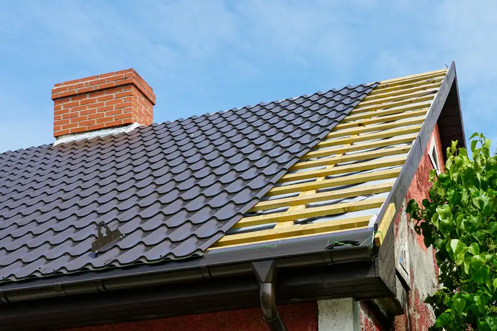 A house roof partially covered with dark metal tiles; the right side shows exposed wooden battens and underlayment. A red brick chimney rises from the roof, installed by a roofer Jasper County, with green foliage along the right edge.
