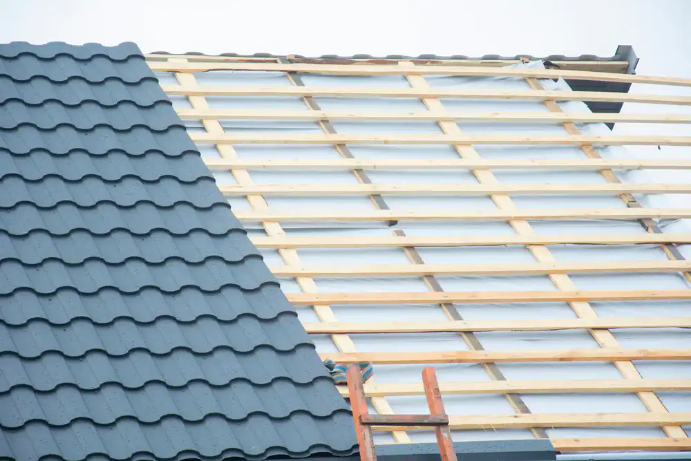 A house roof under construction with one side covered in dark gray metal tiles and the other side showing exposed wooden battens and insulation, with a ladder leaning against the structure.