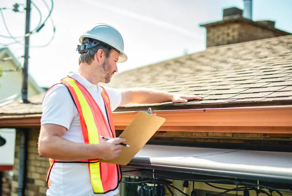 A roofing contractor from Southwest Louisiana, wearing a hard hat and safety vest, examines a house roof while holding a clipboard and pen.