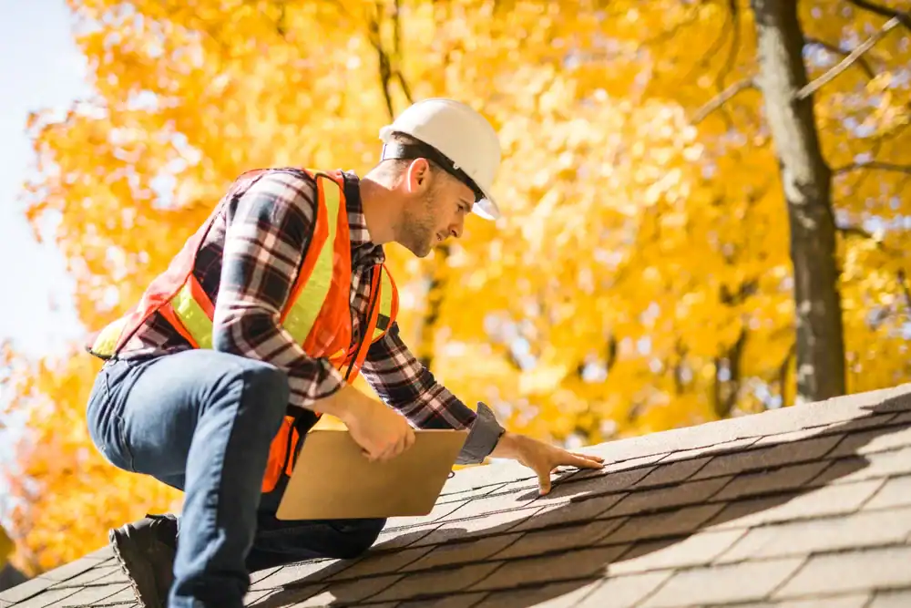A roofing contractor from Southwest Louisiana, wearing a hard hat and safety vest, inspects roof shingles on a house with clipboard in hand. Bright yellow autumn trees are visible in the background.