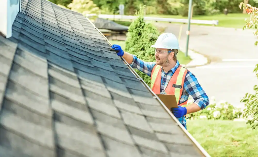 A roofer Jasper County in a hard hat, safety vest, and gloves inspects a house roof with a clipboard on a sunny day, surrounded by greenery and a residential neighborhood.