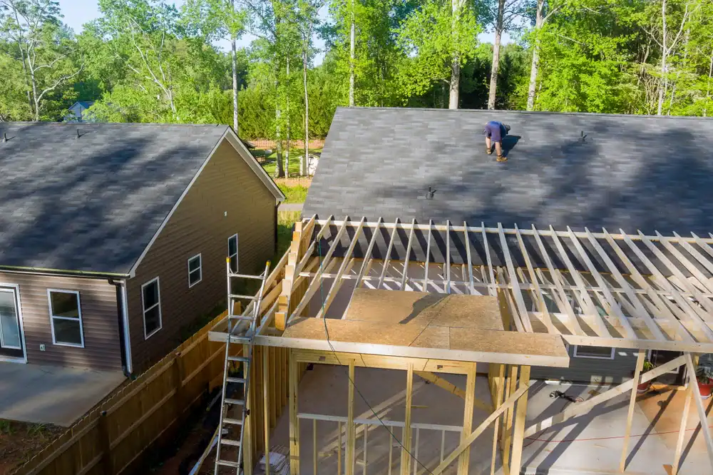 A roofing contractor Southwest Louisiana is working on the roof of a house under construction, with wooden beams and a ladder visible. Another finished house sits nearby, surrounded by trees and greenery in the background.