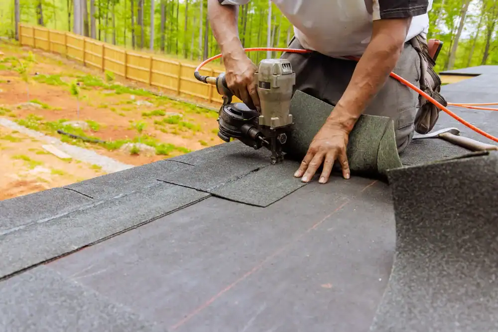 A roofer from Jasper County uses a nail gun to install asphalt roofing shingles on a roof, kneeling and working with both hands, while trees and a wooden fence provide the backdrop.