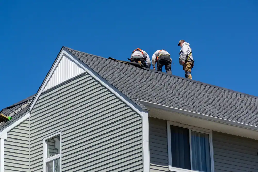 Three workers wearing safety gear, possibly from a roofing contractor Southwest Louisiana, are installing or repairing shingles on a grey house roof under a clear blue sky.