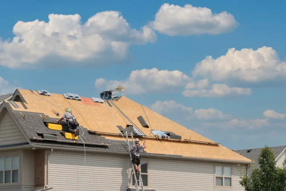Two workers are installing new roofing materials on the roof of a house under a blue sky with scattered clouds. Ladders and equipment are visible, and the roof is partially covered with plywood and roofing sheets.