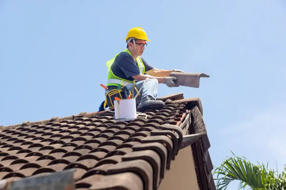 A roofer in Jasper County wearing a yellow hard hat and safety vest repairs a tiled roof, holding a roof tile and sitting near a bucket of tools under a clear blue sky.