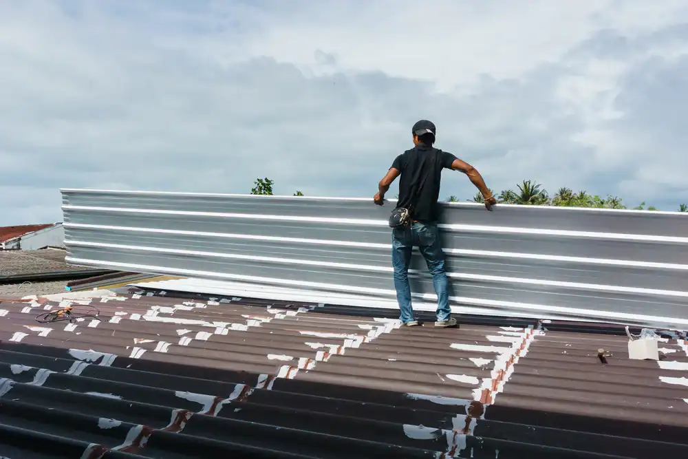 A roofer in Jasper County, TX, wearing a black shirt, jeans, and a cap stands on a rooftop holding a large sheet of metal roofing. The sky is cloudy and trees are visible in the background.