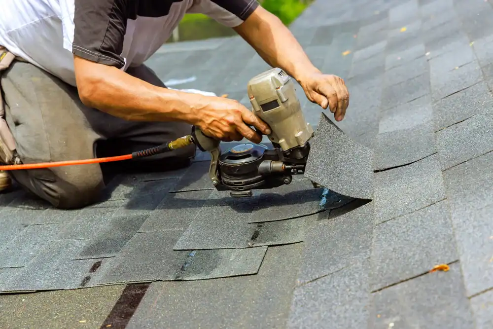 A person kneels on a roof while using a nail gun to install asphalt shingles, securing them in place.