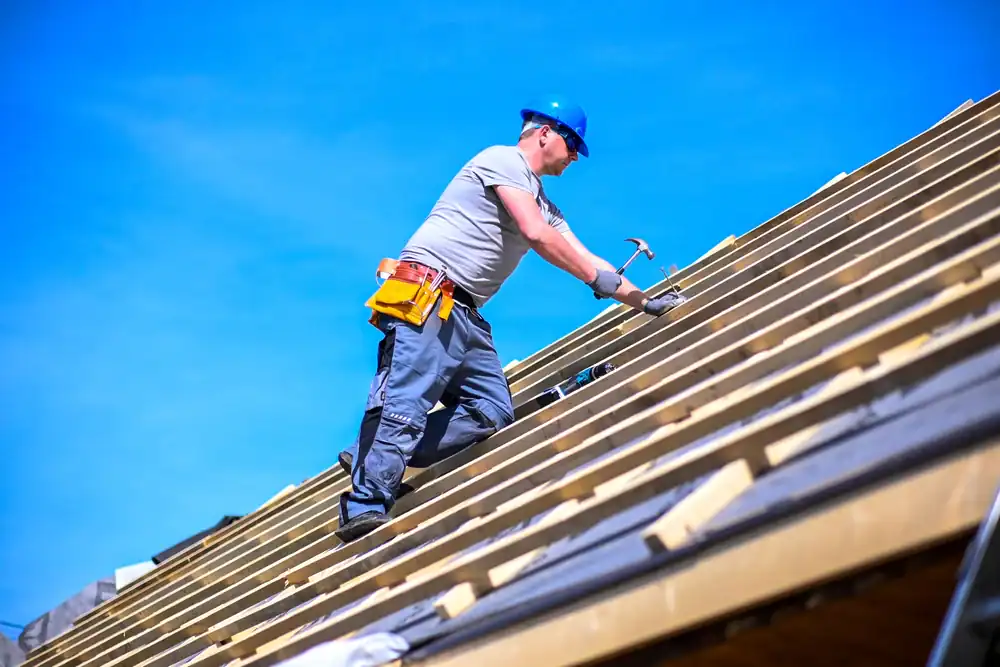 A construction worker wearing a blue hard hat and tool belt uses a hammer while working on a sloped roof under a clear blue sky.