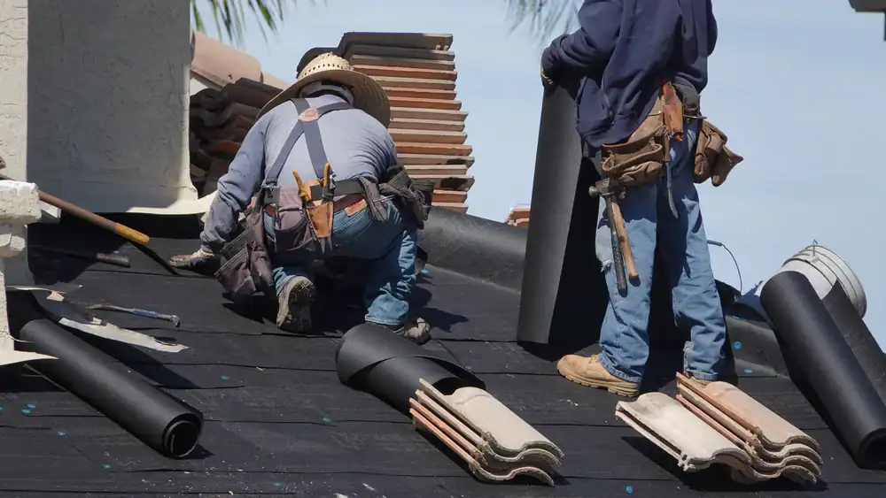 Two workers wearing protective clothing and tool belts are installing roofing materials on a house, with roofing rolls, tiles, and tools spread out around them&mdash;a skilled roofer Jasper County trusts for quality results.