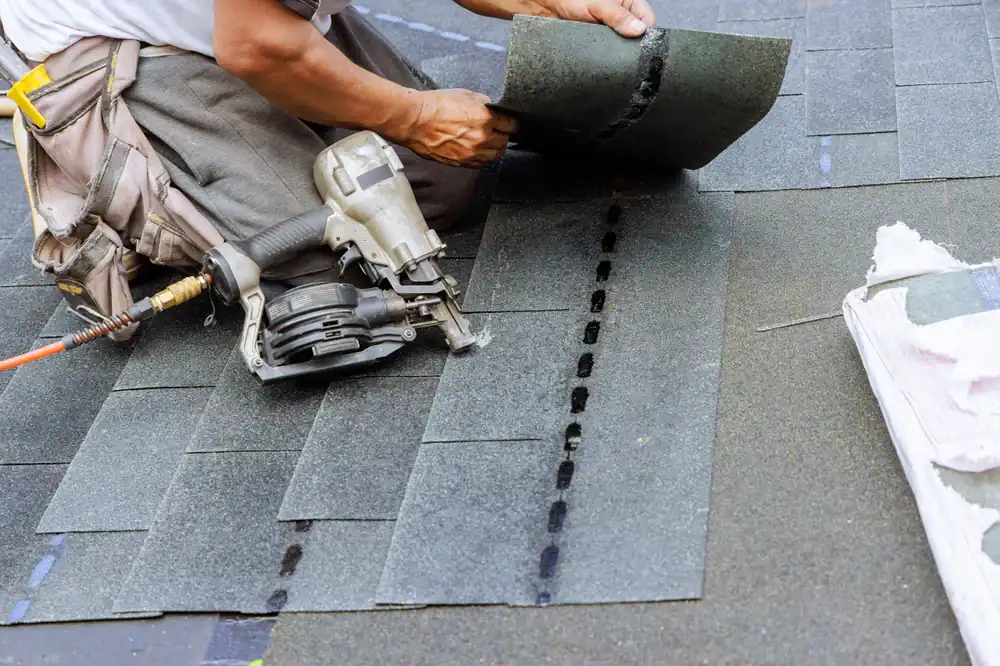 A roofer Jasper County installs asphalt shingles on a roof, using a nail gun and aligning the shingles carefully. A bundle of shingles is visible nearby.