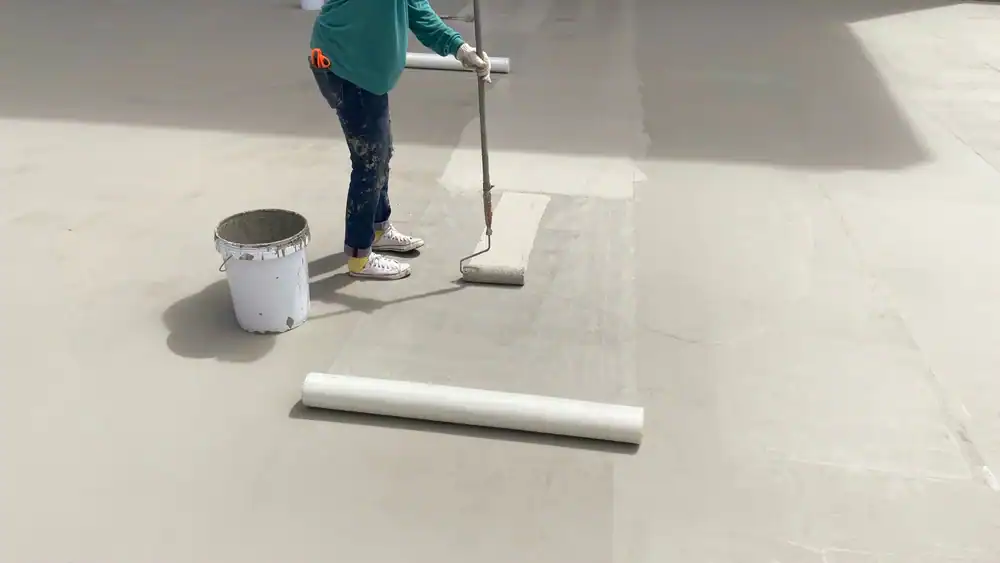 A person uses a paint roller to apply a coating to a concrete floor. A white bucket sits nearby, with another paint roller on the ground. The person, possibly a roofer from Jasper County, wears gloves, jeans, and a green shirt.