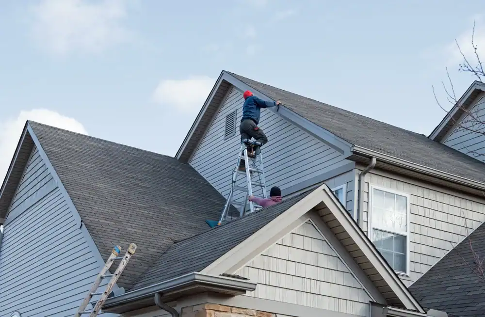 Two people work on the roof of a house; one stands on a ladder while reaching up to the roofline, and the other supports from below. Additional ladders are positioned against the house. The sky is partly cloudy.