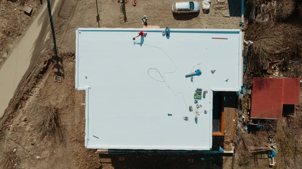 Aerial view of workers installing a white roof membrane on a large rectangular building. Tools and equipment are scattered on the roof, and a car is parked nearby on a dirt area.
