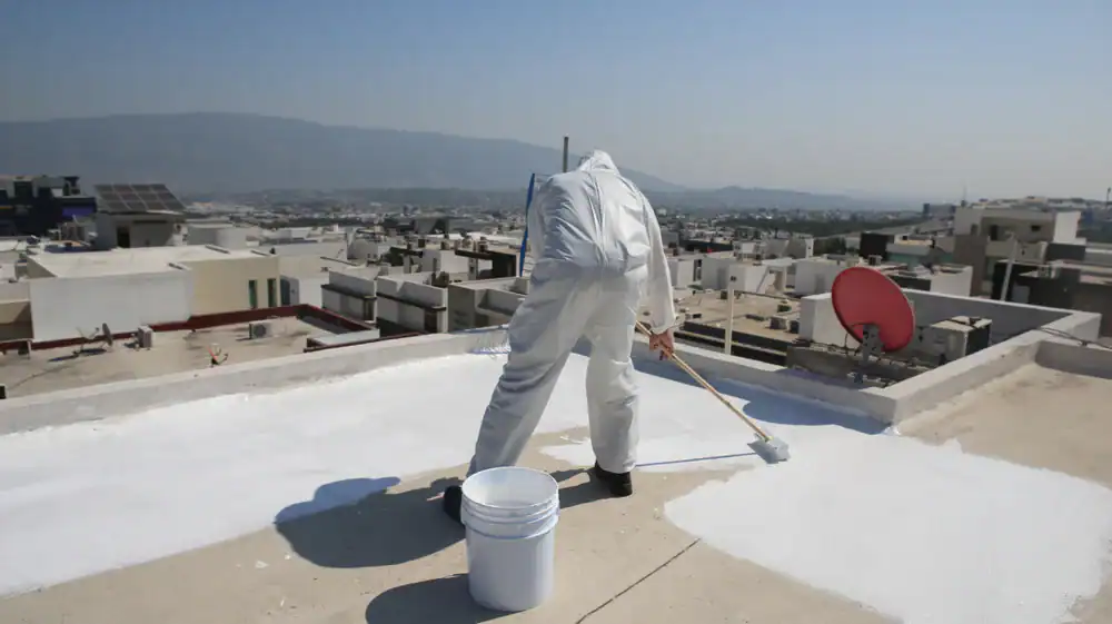 A person in a full white protective suit paints a rooftop white with a roller, overlooking a cityscape with buildings, mountains in the distance, and a red satellite dish nearby.