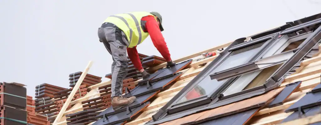 A construction worker in a yellow safety vest and helmet installs roofing materials and windows on a sloped roof, surrounded by wooden beams and stacks of tiles.