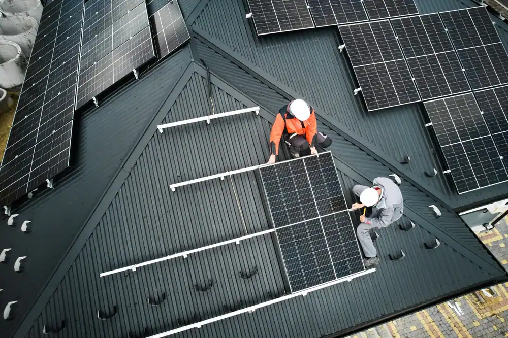 Two workers in safety gear, including a roofer Jasper County expert, install solar panels on the ridged roof of a building. Several panels are already mounted as the team secures another panel in place under the Texas sun.