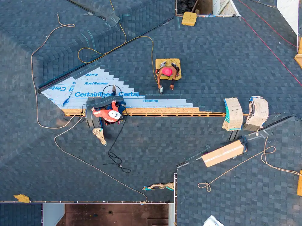 Aerial view of workers installing shingles and roofing materials on a house roof, with tools, cords, and building materials visible around them.