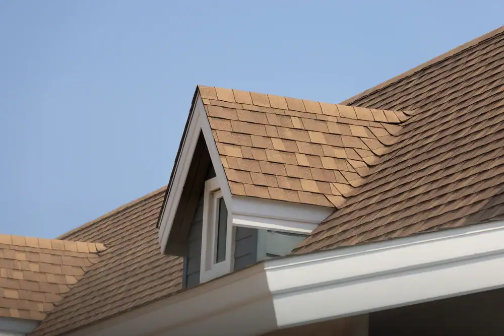 Close-up of a house roof with brown asphalt shingles, a small dormer window, and white trim under a clear blue sky—work by a trusted roofing contractor Southwest Louisiana.