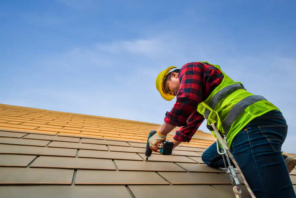 A construction worker in a yellow hard hat and safety vest installs roof tiles with a power drill on a slanted roof under a clear blue sky.