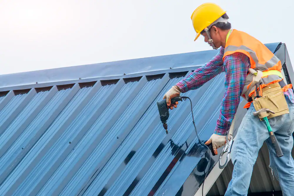 A roofing contractor Southwest Louisiana, wearing a hard hat and safety gear, uses a power drill to install or repair a metal roof on a building.