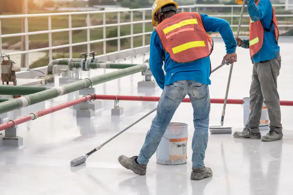 Two construction workers in safety vests and helmets apply a waterproof coating to a flat roof using paint rollers, showcasing the skill of a professional roofer Jasper County. Pipes and railings are visible in the background.