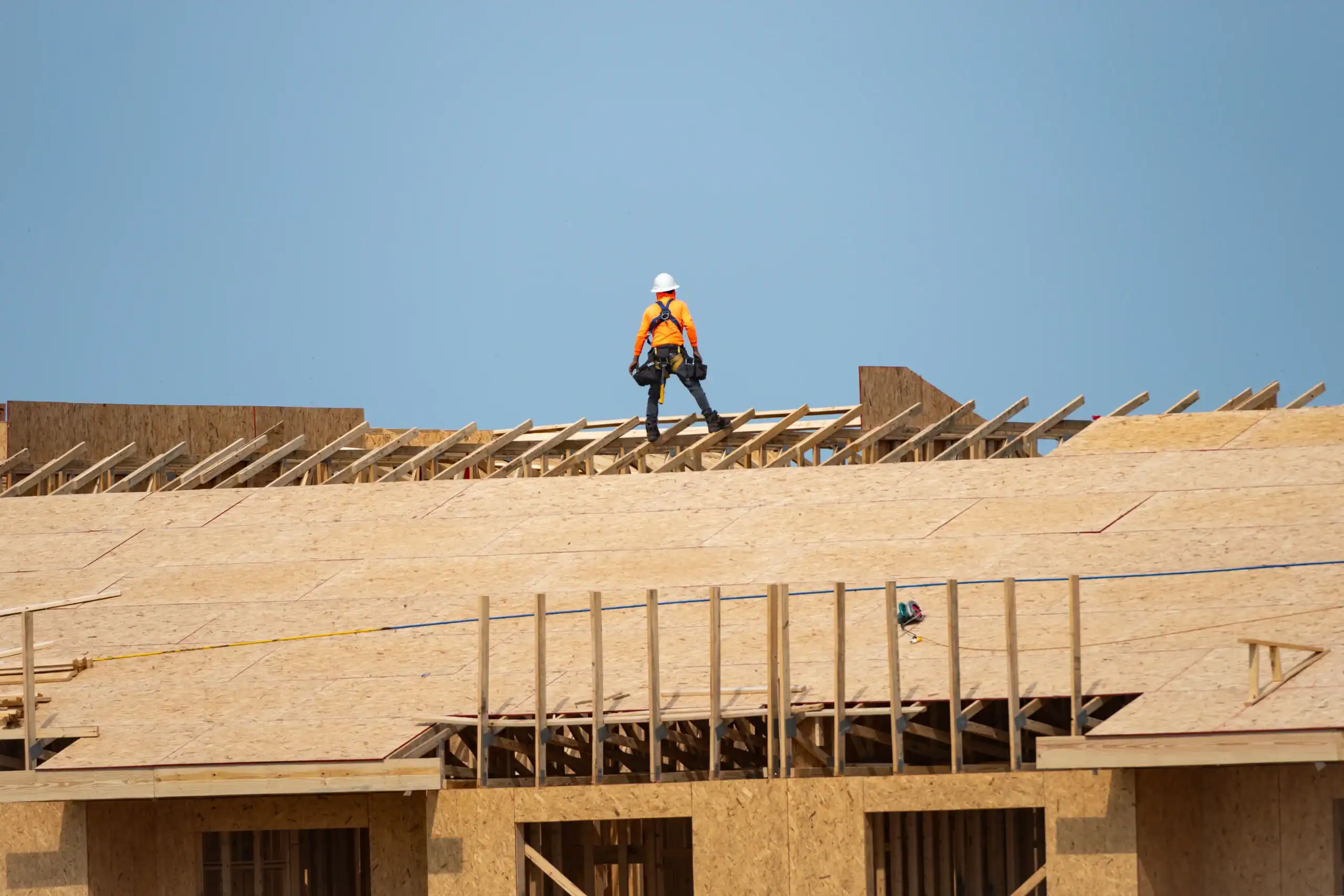 A construction worker in a safety vest and helmet walks on the wooden roof framework of a building under construction, with a clear blue sky in the background.