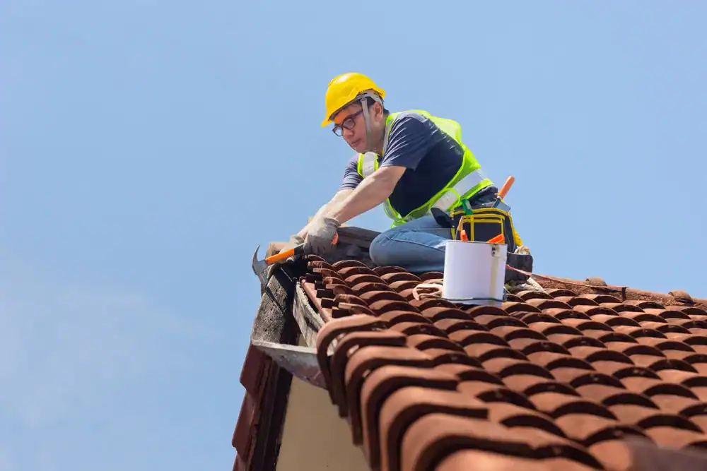 A construction worker in a yellow hard hat and safety vest repairs the edge of a tiled roof with tools and a paint bucket nearby under a clear blue sky.