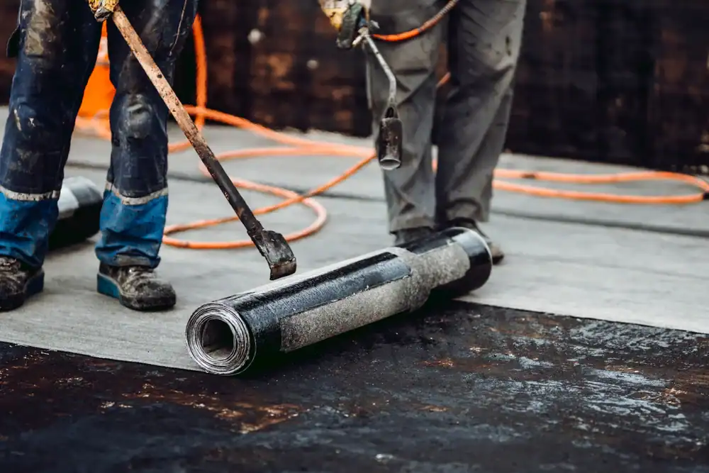 Two workers use a torch to apply heat to a rolled sheet of roofing material on a flat rooftop during construction or repair. Orange cords and tools are visible around them.