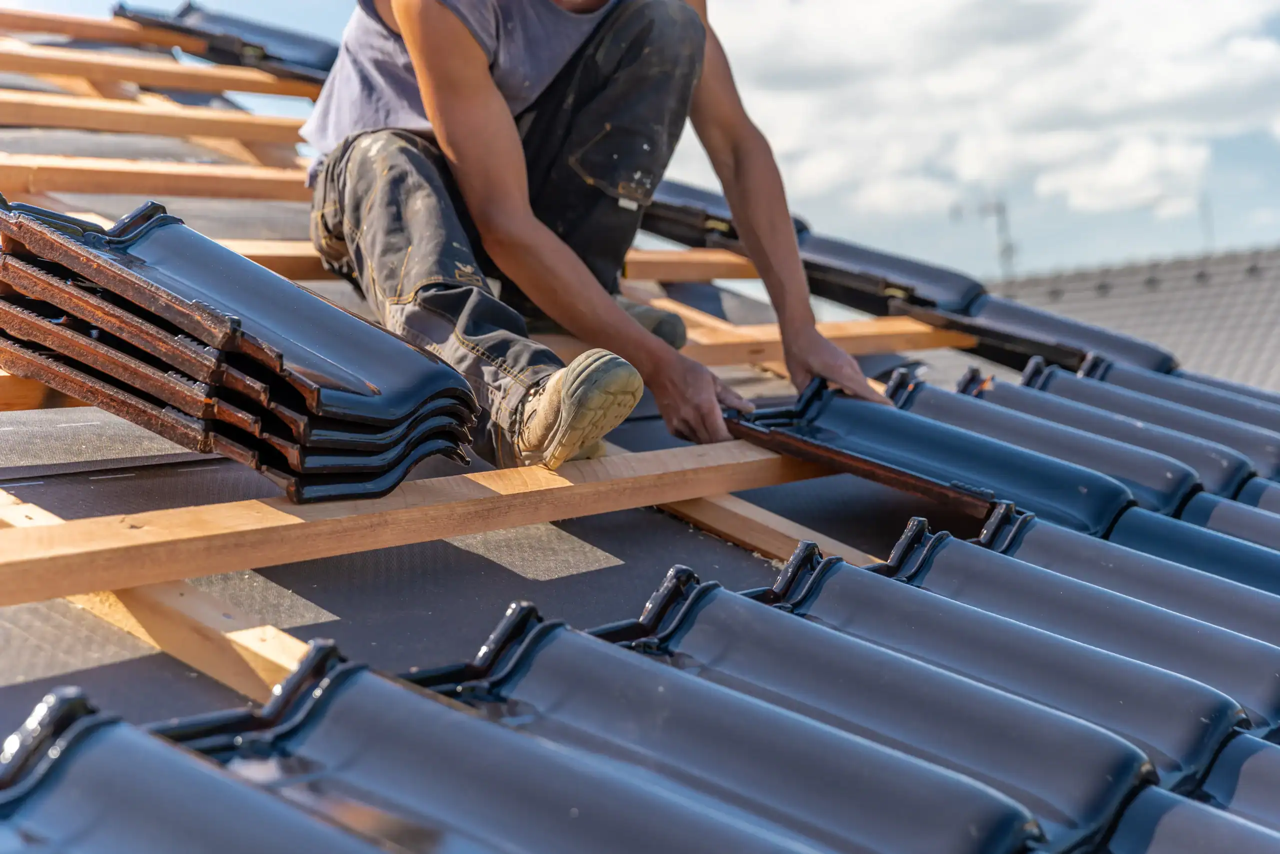 A worker installs shiny black roof tiles on a wooden frame of a sloped roof under a bright sky, with stacks of tiles beside him.