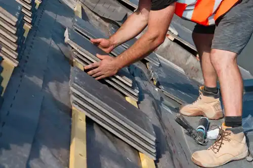 A construction worker wearing boots and an orange vest installs rectangular roof tiles on a building, using both hands to place the tiles accurately on wooden battens. Tools are visible nearby on the roof.