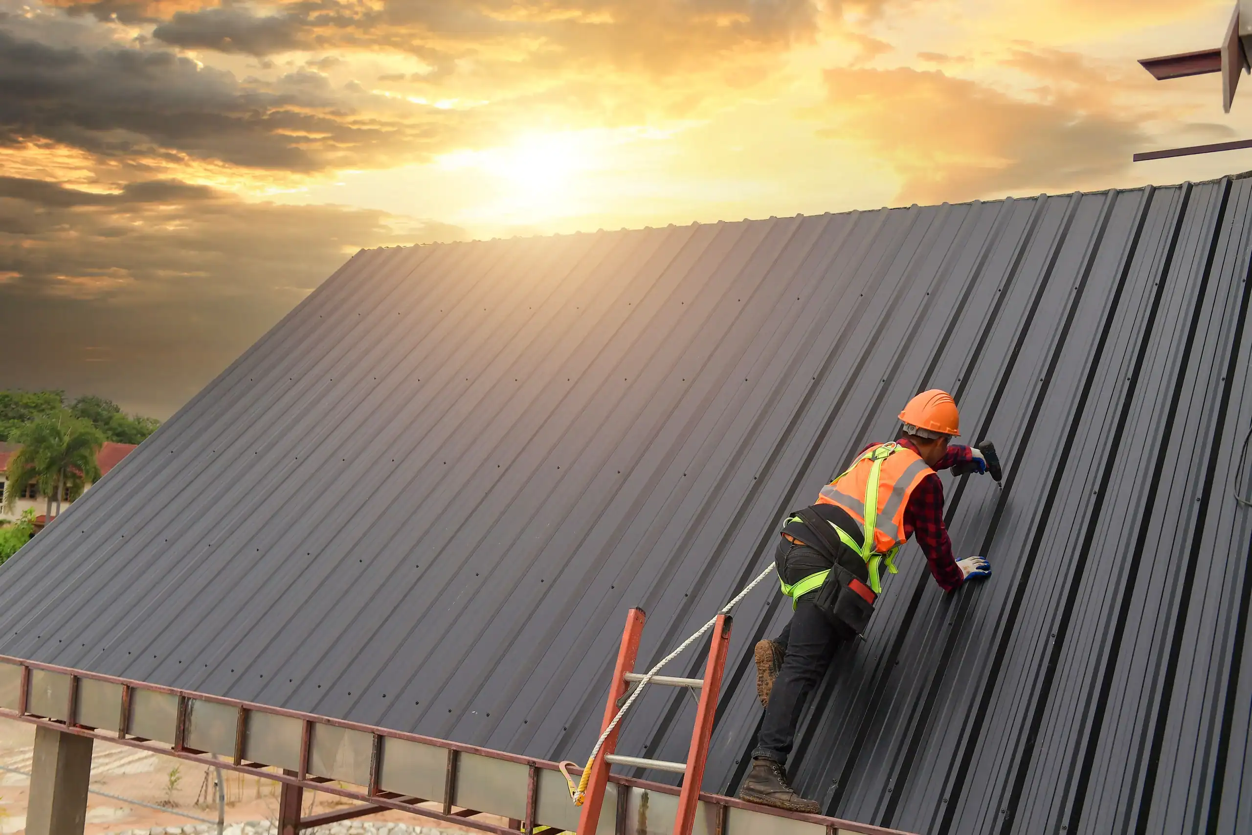 A construction worker wearing safety gear and a harness installs or inspects a metal roof at sunset, with dramatic clouds and sunlight in the background.